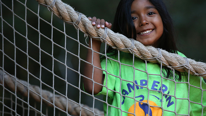 Young girl smiling on a rope bridge wearing a green camp erin shirt