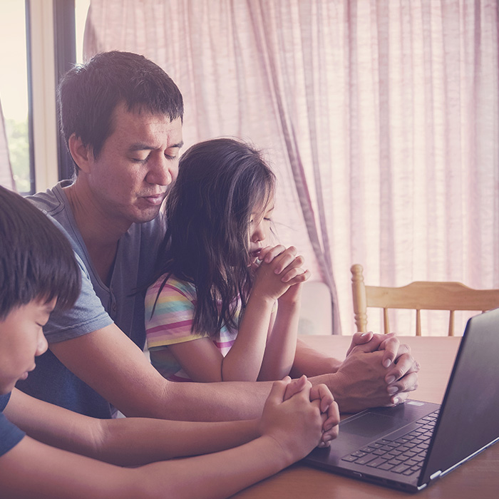 Dad and two children praying in front of a laptop